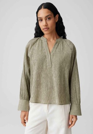 Frau mit langen dunklen Haaren trägt eine strukturierte olivgrüne Bluse mit langen Ärmeln und weiten Manschetten sowie weiße Hosen vor schlichtem Hintergrund.