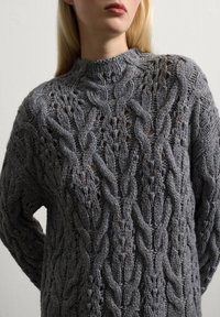 Femme portant un épais pull en tricot gris avec un col montant, présentant des motifs en tresses et ajourés, se tenant devant un fond clair.