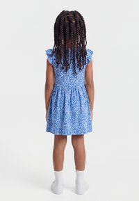 Κορίτσι με μακριά πλεγμένα κοτσίδια στέκεται γυρισμένο με την πλάτη, φορώντας ένα κοντό μπλε φόρεμα με λευκό λουλουδάτο σχέδιο, λευκές κάλτσες και λευκά παπούτσια.