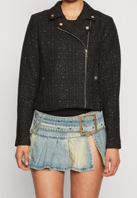 Czarna żakiet tweedowy z metalowymi akcentami, asymetrycznym zamkiem błyskawicznym i klapami, w połączeniu z jasnoniebieską, plisowaną spódnicą dżinsową z szerokim paskiem.