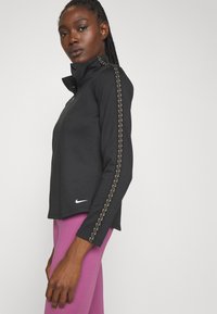 Zwarte sporttop met een hoge kraag, voorzien van gouden stiksels langs de mouwen. De stof is glad en wordt gecombineerd met aansluitende roze leggings.