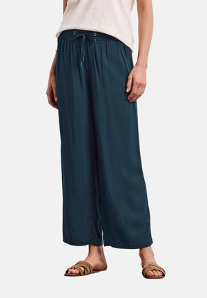 Person iført mørkegrønne vidde bukser med snøre i taljen, hvid ærmeløs top og brune slidesandaler, stående på hvid baggrund.