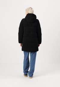 Manteau noir rembourré avec une capuche, présentant un design matelassé. Porté avec un pantalon bleu à motifs et des chaussures beiges, vu de dos.