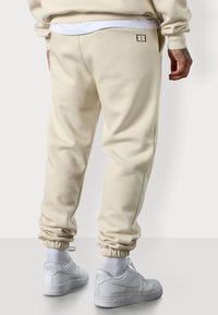 Beige Jogginghosen mit elastischen Bündchen und einem Logo auf der Rückseite. Hergestellt aus weichem Material, mit entspannter Passform und Seitennähten.