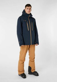 Giacca da sci blu navy con zip dorate e tasche sul petto, abbinata a pantaloni da sci beige e scarponi da sci neri. Il materiale sembra resistente all'acqua e durevole.