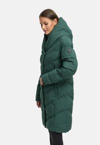 Grön puffjacka med överdimensionerad huva, quiltat mönster och svart logotypknapp på ärmen. Har tryckknappsstängning och sidofickor.