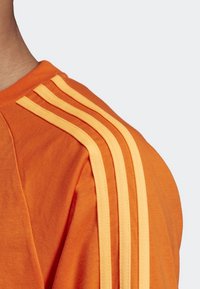 Camiseta naranja con cuello redondo, que presenta tres franjas amarillas a lo largo del hombro. Fabricada en algodón suave, tiene una textura lisa.