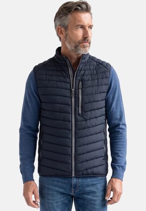 Mann trägt eine marineblaue gesteppte Weste über einem blauen Langarmshirt und blauen Jeans, schaut seitlich vor einem schlichten Hintergrund.