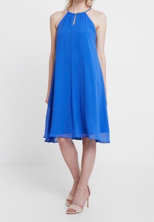 Kvinde iført en ærmeløs, knælang royalblå halterkjole med nøglehul-halsudskæring og beige højhælede sandaler, foran en hvid baggrund.