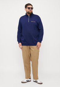 Sudadera con cremallera de color marino y cuello alto, con texto bordado en dorado. Combinada con pantalones caqui claros y zapatillas blancas y negras.