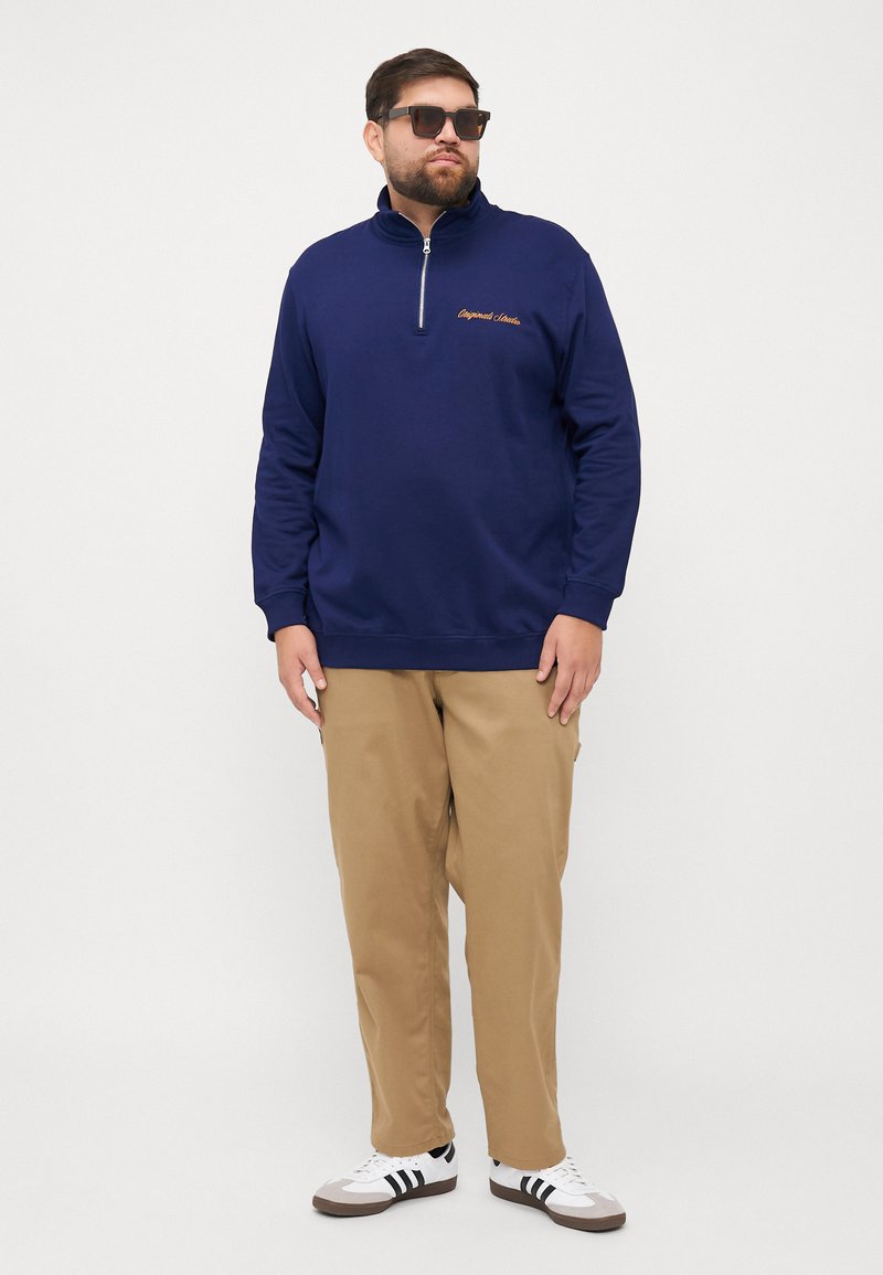 Marine blauwe rits-sweater met een hoge kraag, voorzien van goud geborduurde tekst. Gekleed met licht kaki broek en zwart-witte sneakers.