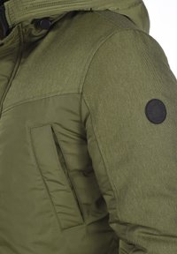 Chaqueta verde oliva con un panel de hombro texturizado, cierre de cremallera y un logotipo negro redondo en la manga. Cuenta con un bolsillo lateral.