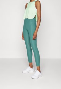 Vihreä hihooton crop-top, jossa on korkea kaula-aukko, yhdistettynä teal-värisiin legginsseihin. Valkoiset lenkkarit, joissa on teksturoitua yksityiskohtaa, täydentävät asusteen.