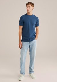 Tricou de culoare albastru închis, cu mânecă scurtă, dintr-un material neted, asortat cu blugi din denim de culoare albastru deschis. Modelul stă cu o mână în buzunar, purtând adidași albi.