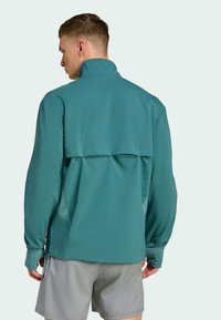 Veste de sport teal avec col montant, manches longues et détail d'ouverture à l'arrière. Fabriquée en matériau léger et respirant. Associée à des shorts gris.