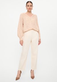 Blusa in pesco color chiaro con motivo ondulato testurizzato, scollatura a V e maniche a palloncino abbinata a pantaloni beige su misura e tacchi marroni.