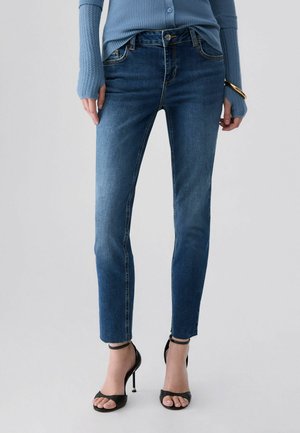 Woman wearing blue slim-fit jeans, blue ribbed sweater, gold bracelet, and black high-heeled sandals standing against a plain background.