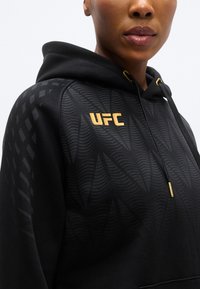Sweat à capuche noir avec un design texturé présentant des motifs d'ondes courbes, des accents dorés et un logo UFC jaune bien visible. Cordon de serrage ajustable inclus.