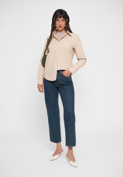 Light beige button-up shirt, long sleeves, paired with dark blue straight-leg jeans and white mules. Features subtle stitching details.
