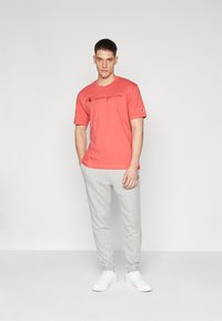 T-shirt corail à manches courtes avec logo "Champion" brodé, associé à un pantalon de jogging gris clair et des baskets blanches, sur un fond uni.