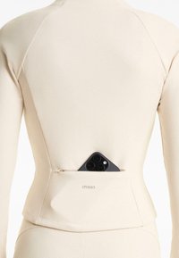 Top a maniche lunghe a coste beige con tasca sul retro, caratterizzato da una cerniera e uno smartphone nero parzialmente visibile. Logo "OYSHO" in vita.