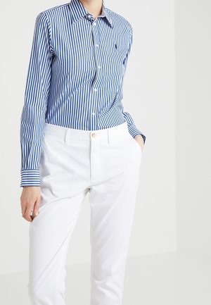 Persoon draagt een blauw-wit verticaal gestreept overhemd, ingestopt in een witte broek, met één hand in de zak.