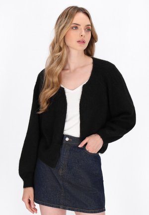 Frau mit langen blonden Haaren, die eine schwarze, flauschige Strickjacke, ein weißes Oberteil und einen dunkelblauen Jeansrock trägt, posiert mit einer Hand in der Tasche.