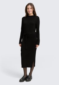 Mujer de pie vistiendo un vestido midi negro de manga larga con cinturilla, medias negras y mocasines negros frente a un fondo claro y liso.