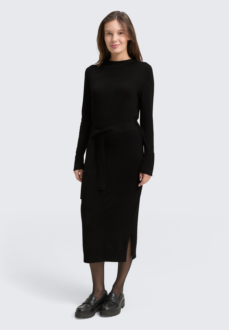 Mujer de pie vistiendo un vestido midi negro de manga larga con cinturilla, medias negras y mocasines negros frente a un fondo claro y liso.