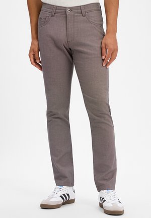 Man met slim-fit bruine broek en witte sneakers met zwarte strepen, staand tegen een witte achtergrond.