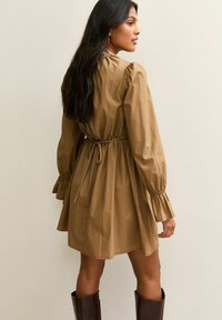 Donna con lunghi capelli scuri che indossa un vestito beige a maniche lunghe con cintura in vita e stivali marroni fino al ginocchio, in piedi contro uno sfondo semplice.