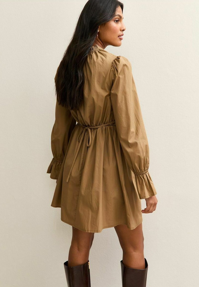 Donna con lunghi capelli scuri che indossa un vestito beige a maniche lunghe con cintura in vita e stivali marroni fino al ginocchio, in piedi contro uno sfondo semplice.