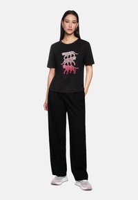 Black t-shirt with colorful graphic of stylized cats in pink and white, paired with loose black trousers and light athletic shoes.