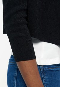 Άτομο που φοράει μαύρο πουλόβερ με μακριά μανίκια πάνω από ένα λευκό πουκάμισο, συνδυασμένο με μπλε τζιν τζιν, δείχνοντας ένα κοντινό πλάνο του βραχίονα και της περιοχής της μέσης.