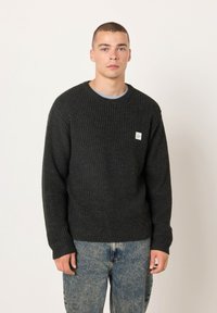 Tamnosivi rebrasti pulover od pletenine s dugim rukavima i okruglim dekolteom, s malim bijelim natpisom na prsima. Ležerni kroj.