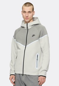 Sudadera con cremallera de Nike en gris claro con detalles en gris más oscuro. Presenta una capucha, bolsillos laterales con cremallera y un logo en el pecho.