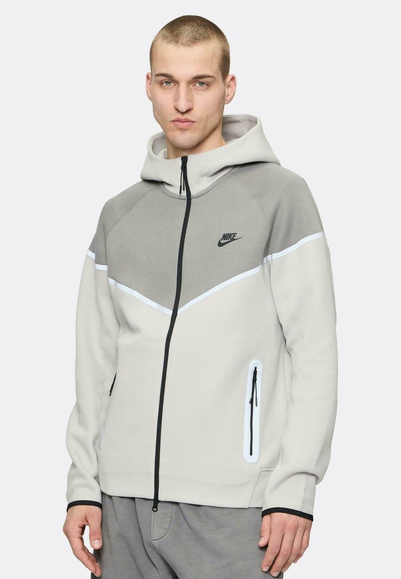 Sudadera con cremallera de Nike en gris claro con detalles en gris más oscuro. Presenta una capucha, bolsillos laterales con cremallera y un logo en el pecho.