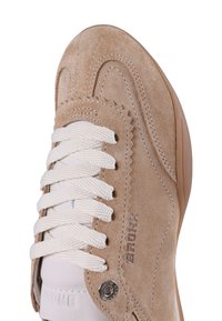 Beige suedesneakersit, jossa on valkoiset nahkaosuudet, paksut pyöreät nauhat ja pyöreä kärki. Siinä on ommeltuja yksityiskohtia ja logo sivulla.