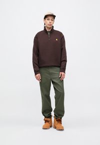 Polaire marron à fermeture éclair quart, pantalon décontracté vert, et bottes de randonnée couleur tan. Présente un détail de logo sur la poitrine et un tissu texturé.