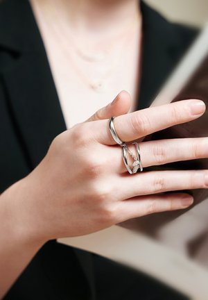 To sølv ringe bæres på en hånd, med en glat ring og en anden med et snoet design og stenaccenter. Glat hudtekstur er synlig.