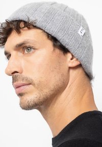 Graue gerippte Strickmütze mit weicher Textur, versehen mit einem kleinen weißen Etikett. Das Gesicht des Modells ist teilweise sichtbar und zeigt den natürlichen Hautton und Details.