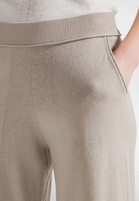 Lähikuva beigeistä neulehousuista, joissa on resorivyötärö ja sivutasku, käsi osittain taskussa.