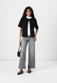 Black short-sleeve top with a split front, layered over a white shirt. Wide-leg grey jeans with a faded texture. Black handbag and patterned flats.