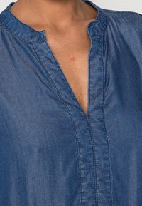 Denimskjorta med V-ringning, vertikal sömnad fram och en lätt glans på ett medium blått tyg. Slät textur.