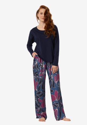 Kobieta o długich rudych włosach, ubrana w granatową bluzkę z długim rękawem i luźne niebieskie spodnie z dużym różowo-czarnym kwiatowym wzorem, stojąca boso.