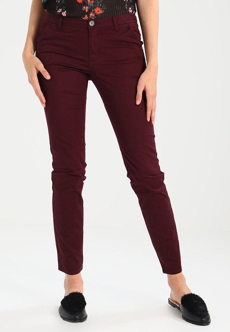 Bordeauxkleurige, aansluitende broek met een gladde textuur, voorzien van een knoopsluiting en zijzakken, gecombineerd met zwarte, zachte instapschoenen.