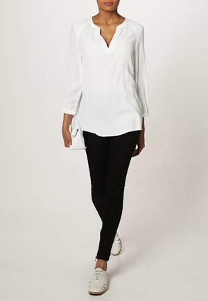 Hvid langærmet bluse med V-hals, der har subtile samlinger ved skuldrene, parret med sorte slim-fit bukser og hvide snørebåndssko.