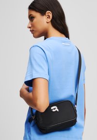 Black rectangular crossbody bag made of durable material with a silver hardware accent. Features a shoulder strap and subtle branding detail.