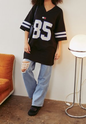 Schwarzes übergroßes Trikot mit weißen und blauen Akzenten, das die große Zahl "85" und ein Logo zeigt. Kombiniert mit hellblauen, zerrissenen Jeans und schwarzen Schuhen.