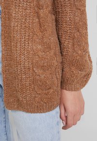 Brown knitted cardigan with a textured pattern, featuring ribbed cuffs and hem. Paired with light blue denim jeans for contrast.